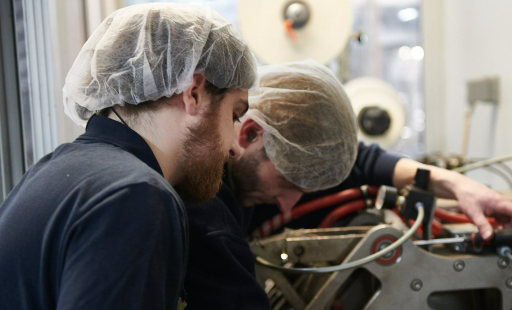 Two technicians in hair nets work collaboratively on a complex machine in a modern engineering workspace.