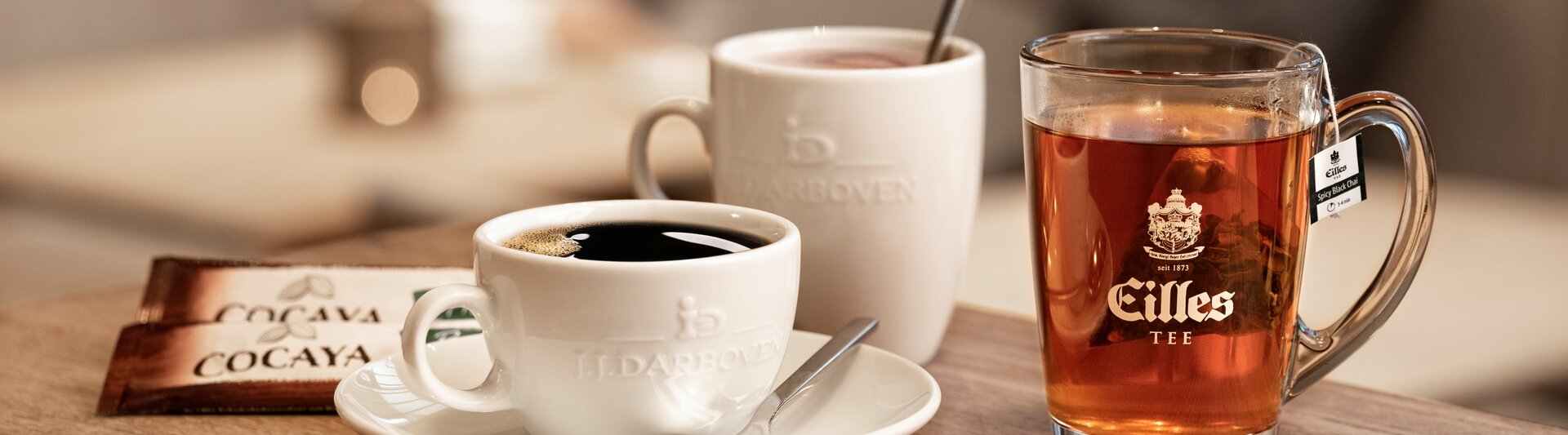 A black coffee and a glass of tea sit on a wooden table, with two people chatting in the blurred background.