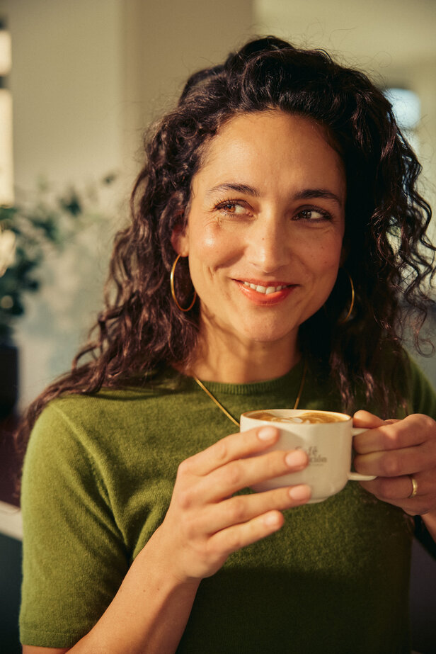 A woman enjoys a coffee in a café, radiating warmth and joy while holding her cup with a smile.