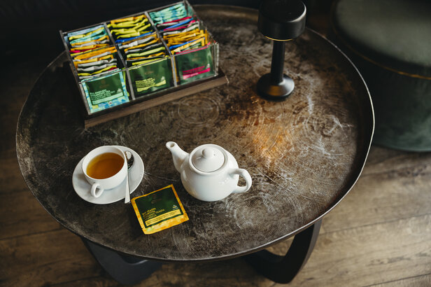 Eine weiße Teekanne und eine Tasse Tee stehen auf einem rustikalen Holztisch mit verschiedenen Teebeuteln im Hintergrund.