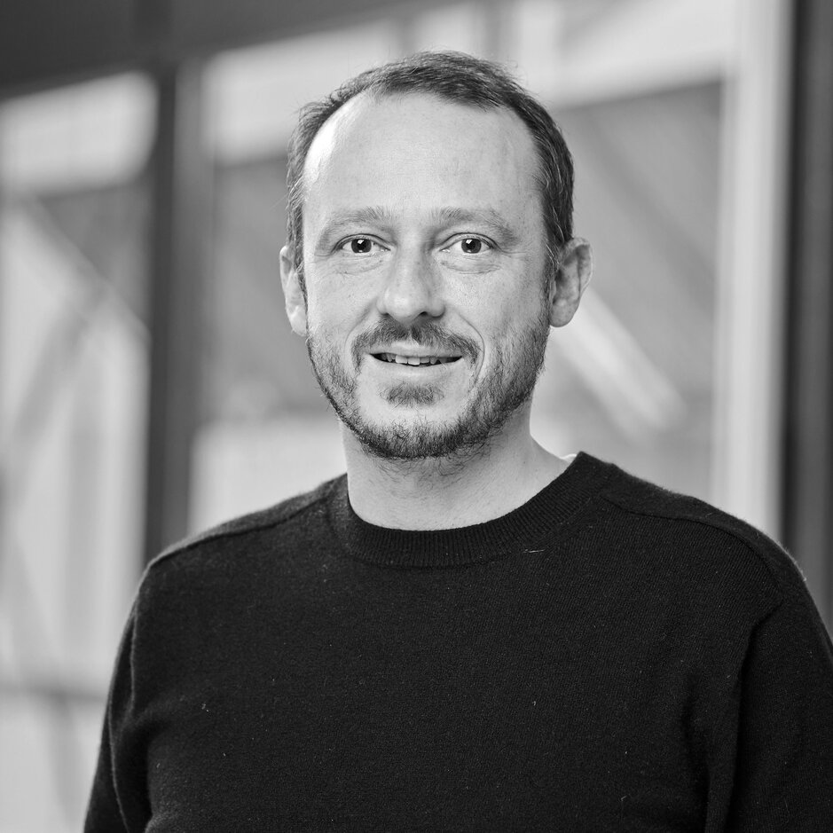 Porträt eines Mannes, der einen schwarzen Pullover trägt und in einer modernen Büroumgebung mit natürlichem Licht lächelt.