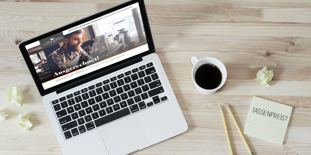Laptop auf einem Holztisch, auf dem eine Website mit Kaffeepreisen angezeigt wird, daneben eine Kaffeetasse und ein Notizblock mit der Aufschrift "TASSENPREIS?".