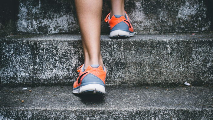 A person ascends concrete stairs wearing vibrant orange and grey sports shoes, symbolising resilience and effort.