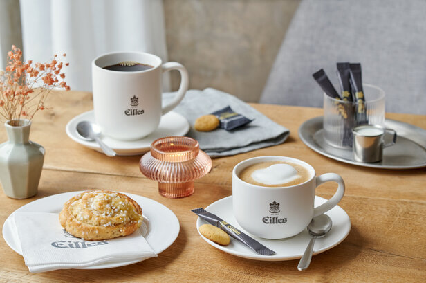 Eilles-Kaffeebecher mit einem Keks und Blumen auf einem Holztisch, die eine warme, einladende Café-Atmosphäre schaffen.