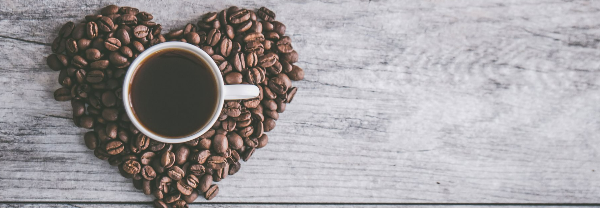 Ein herzförmiges Arrangement aus Kaffeebohnen umgibt eine Tasse mit dunklem Kaffee auf einem Holztisch.