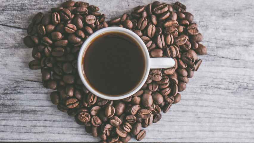 Ein herzförmiges Arrangement aus Kaffeebohnen umgibt eine Tasse mit dunklem Kaffee auf einem Holztisch.