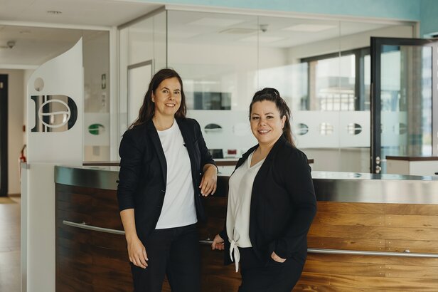 Zwei berufstätige Frauen stehen selbstbewusst an der Rezeption eines modernen Büros und präsentieren ein einladendes Geschäftsumfeld.