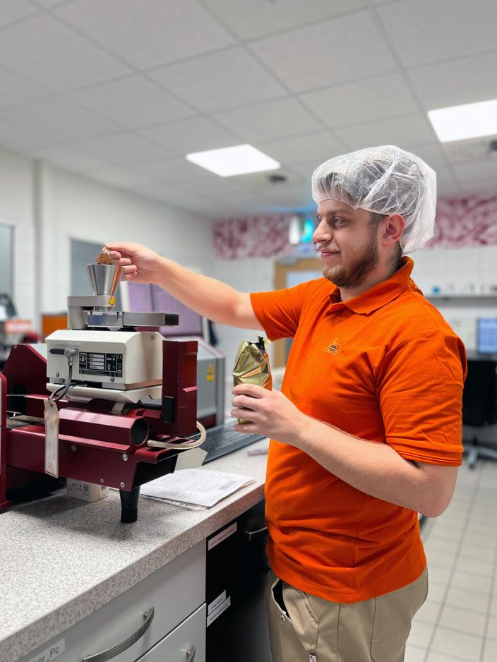 Labortechniker beim Wiegen von Zutaten in einem Qualitätskontrolllabor. Er trägt ein Haarnetz und eine orangefarbene Uniform, um die Lebensmittelsicherheit zu gewährleisten.