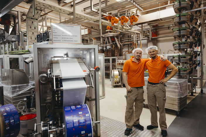 Zwei Fabrikarbeiter in orangefarbenen Hemden posieren stolz in einer modernen Verpackungsanlage mit Maschinen im Hintergrund.