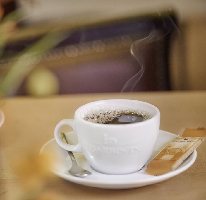Steaming coffee in a white cup on a rustic table, complemented by a sweetener packet and blurred floral background.