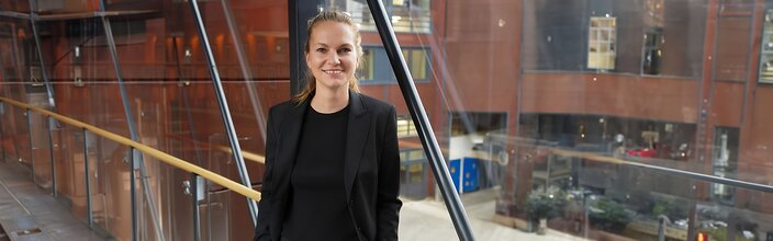 Business professional in a sleek black blazer, standing confidently in a modern office environment with glass architecture.