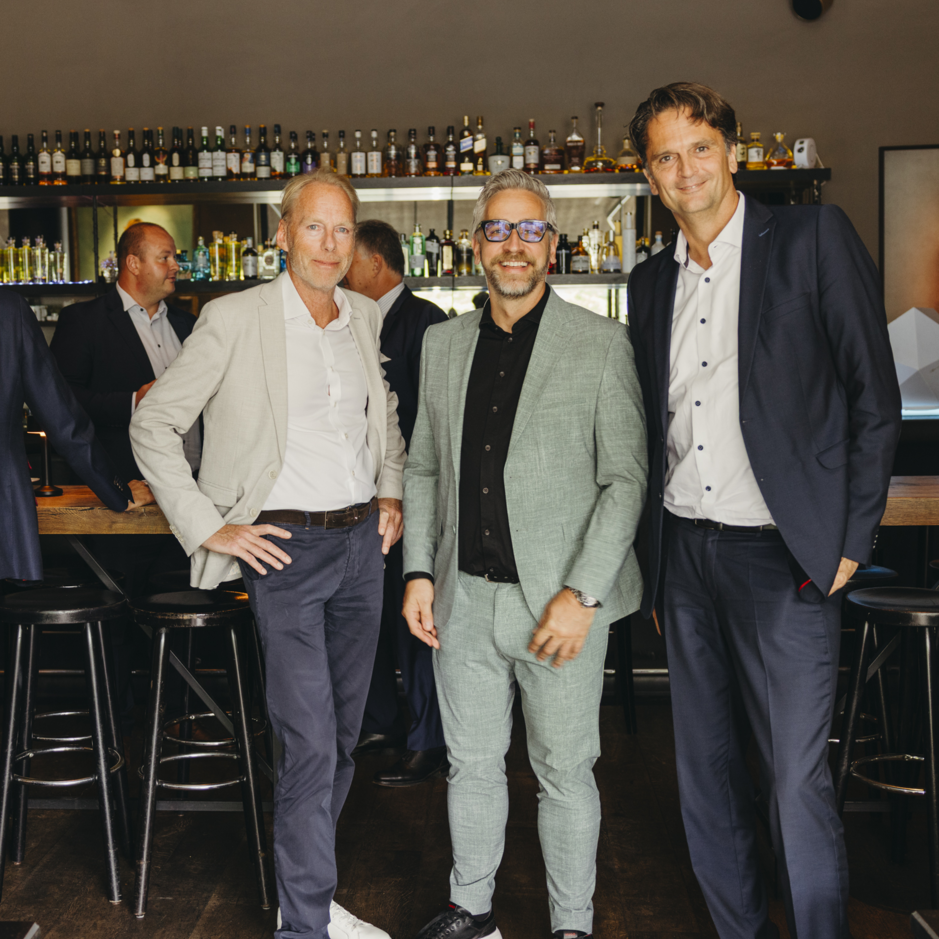 Drei Männer in eleganter Kleidung posieren in einer stilvollen Bar, mit Flaschenregalen im Hintergrund und Kollegen, die sich unterhalten.