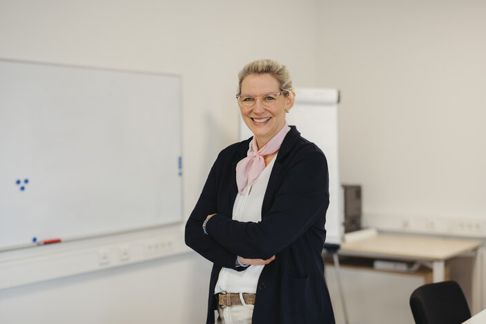 Lächelnde professionelle Frau mit Brille posiert selbstbewusst in einem hellen Büro mit einem Whiteboard im Hintergrund.