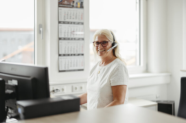 Smiling woman with glasses and headset at her desk, working in a bright office environment.