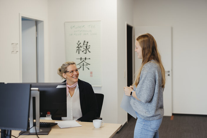 A professional woman smiles at a colleague in an office, promoting a collaborative workplace atmosphere.