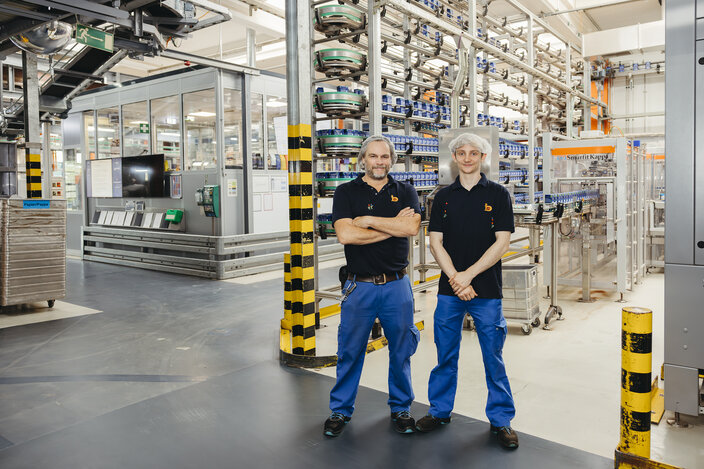 Zwei Fabrikarbeiter posieren selbstbewusst in einer modernen Produktionsstätte und zeigen Teamarbeit in einem lebendigen Arbeitsumfeld.