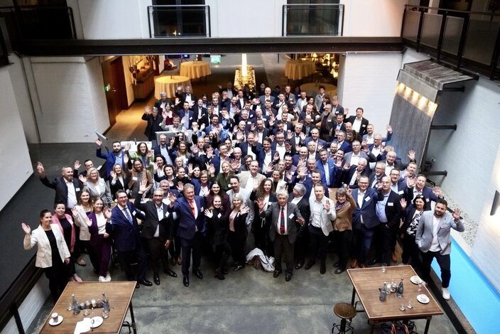 Networking-Veranstaltung mit verschiedenen Fachleuten, die winken und lächeln, in einem modernen Veranstaltungsort mit einer entspannten Atmosphäre.