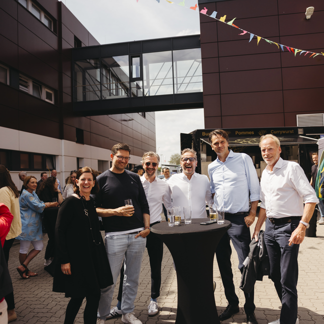 Die Gäste genießen die Getränke bei einer Veranstaltung im Freien, umgeben von Wimpeln und einem modernen Gebäude, das eine festliche Atmosphäre schafft.