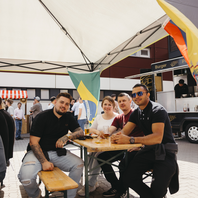Freunde, die auf einem belebten Lebensmittelmarkt unter einem Vordach Getränke genießen, mit einer brasilianischen Flagge im Hintergrund.