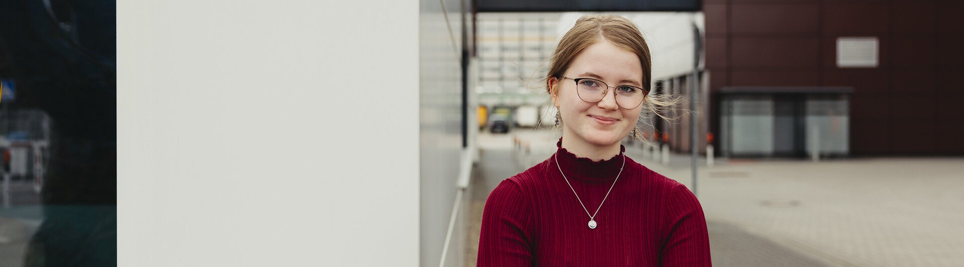 Ein junger Berufstätiger in einem roten Pullover steht selbstbewusst im Freien und zeigt moderne Architektur im Hintergrund.