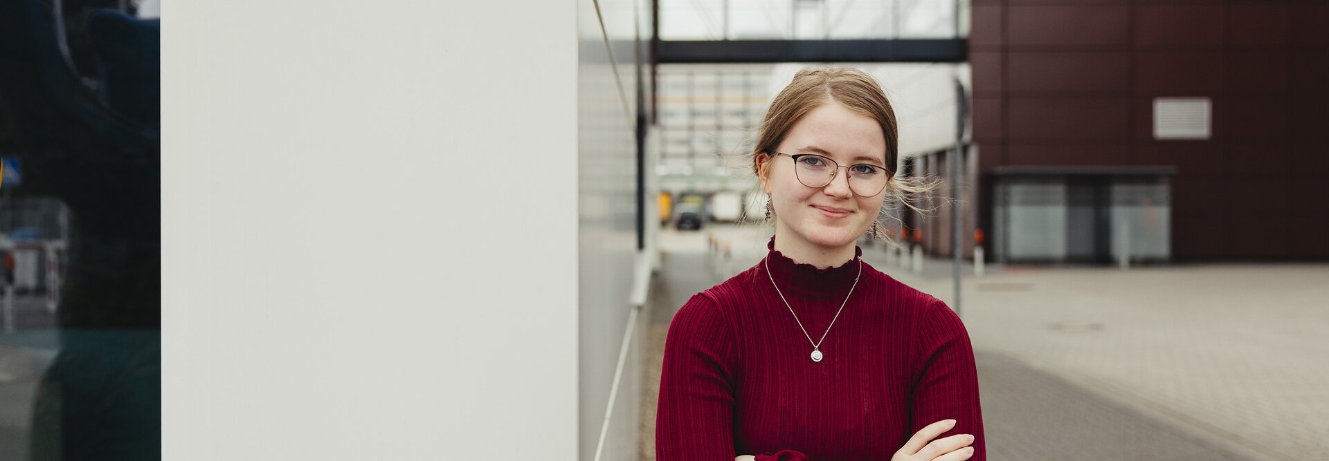 Ein junger Berufstätiger in einem roten Pullover steht selbstbewusst im Freien und zeigt moderne Architektur im Hintergrund.