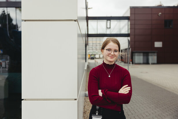 Ein junger Berufstätiger in einem roten Pullover steht selbstbewusst im Freien und zeigt moderne Architektur im Hintergrund.