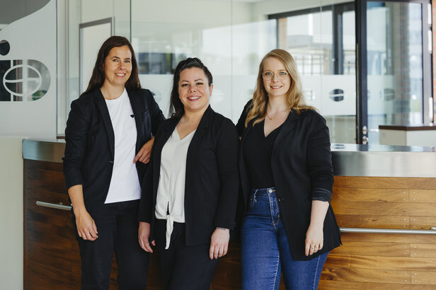 Three professional women pose together at a modern office reception area, showcasing confidence and teamwork.