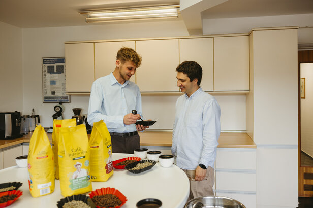 Zwei Männer diskutieren über Kaffeeverkostung in einer modernen Küche, umgeben von Tüten mit Kaffee und Probierbechern.